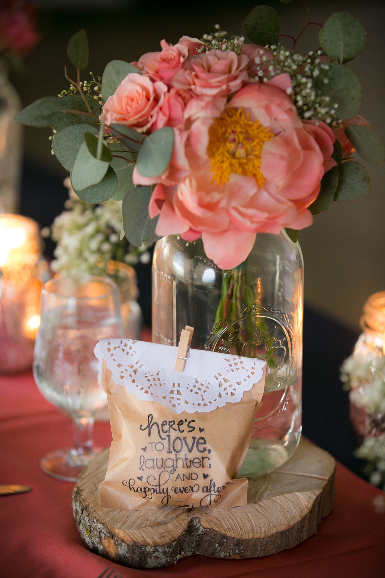 A table center piece at a wedding. Flowers in a glass jar standing on a small cut of natural looking wood, and a small gift bag in front of it.